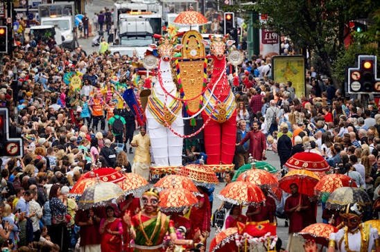 Giant Kerala style puppets and red umbrellas made for the 2019 Manchester Day Parade, carried through a crowded street.